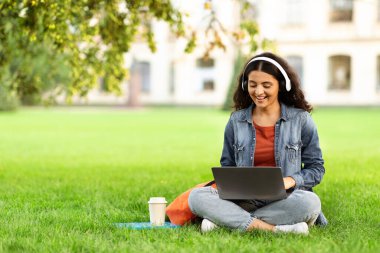 Yeşil çimenlikte dizüstü bilgisayarla oturan mutlu Doğulu Bayan Öğrenci, Park 'ta bilgisayar kullanan güzel Hintli Kadın, Çevrimiçi Eğitim ve Uzaktan Öğrenmenin tadını çıkartıyor, Boşluğu Kopyala