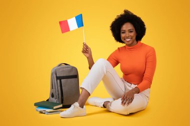 Sırtında sırt çantası ve kitaplarıyla sarı bir arka planda çapraz bacaklı bir Fransız bayrağı taşıyan kıvırcık saçlı, binlerce yıllık gülümseyen Afrikalı Amerikalı kadın.
