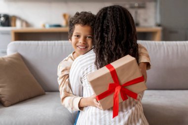Annesine kırmızı kurdeleli sürpriz hediyesini sunan küçük siyah çocuk, annesine sarılırken, sevgi dolu oğlu, annesini anneleriyle selamlıyor, evde neşe anı yakalıyor.