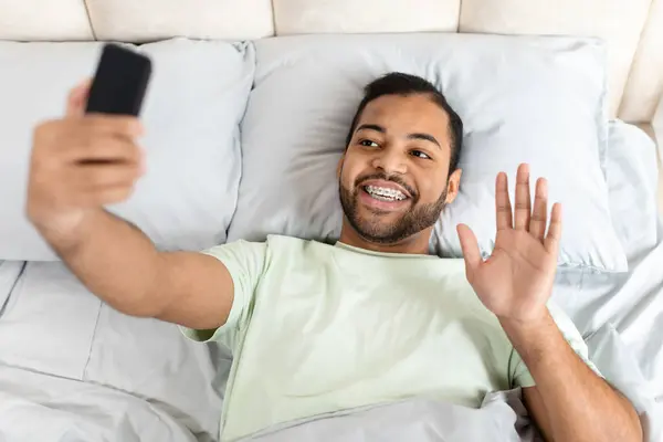 ベッドに横たわっているパジャマを身に着けているハンサムなひげ付きのアフリカ系アメリカ人の男のトップビューは 目覚めた後 ガールフレンドと朝のビデオチャットを持って 携帯電話の画面で振る — ストック写真