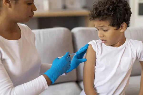 Black doctor woman in protective gloves making vaccination for little boy in home interior, closeup shot of scared african american male kid getting immunization against various disease, closeup