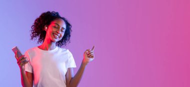 Kıvırcık saçlı gülümseyen genç bir kadın, kulaklıkla müzik dinlemekten zevk alan, akıllı telefon tutan, gradyan mordan pembe arka plana, kopyalama alanı olan bir panorama