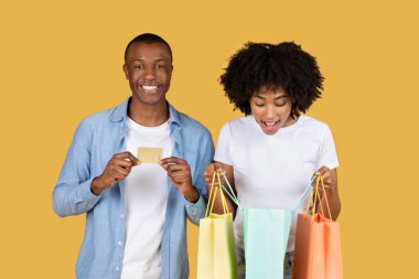 Heyecanlı gülümseyen Afro-Amerikan alışveriş kolikler renkli alışveriş çantaları ve bir kredi kartı ile, başarılı alışverişlerinde mutlu bir şekilde gülümsüyorlar sağlam sarı arka planda, stüdyo