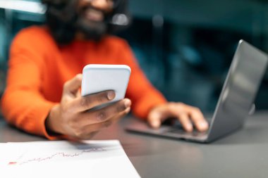 Dijital dünya ve modern iş konsepti. Hintli girişimci karanlık ofiste çalışıyor, akıllı telefonda mobil uygulama kullanıyor ve bilgisayar laptopunda daktilo kullanıyor, kırpılmış