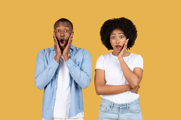 Surprised African American couple with mouths open in shock, the man wearing a blue shirt and the woman in a white tee, against a yellow background, expressing astonishment