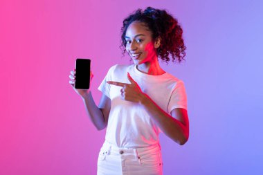 Neşeli siyah kıvırcık saçlı kadın boş akıllı telefon ekranı sunuyor, cihaza işaret ediyor, pembe ve mor gradyan arkaplana karşı, mobil uygulama promosyonunu öneriyor