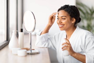 Güzellik rutini. Mutlu Siyah Kadın Evde Aynanın Yakınındaki Cilt Temizleme, Güzel Afrikalı Amerikalı Kadın Makyaj Sütü ve Pamuk Pedi Kullanıyor, Yansımasına Bakıyor, Cildinin Zevkini Çıkartıyor