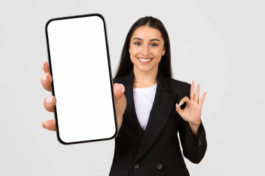 Kendine güvenen genç iş kadını elinde boş ekranlı akıllı telefonu tutuyor ve iyi bir jest yapıyor, iş uygulamasını veya web sitesini hafif arka planda tanıtıyor,