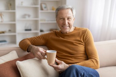 Neşeli gri saçlı, günlük kıyafetler giyen yaşlı bir adam elinde çay fincanıyla kanepede oturuyor ve kameraya gülümsüyor. Pozitif büyükbaba evde sabah kahvesini içiyor, boşluğu kopyalıyor.