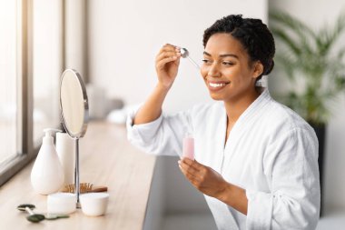 Şişeden yüz serumu uygulamak için gülen zenci kadın, evde güzellik rutini yapan mutlu Afro-Amerikan kadın, rahat banyodaki güzellik ürünleriyle makyaj aynasının karşısında oturuyor.