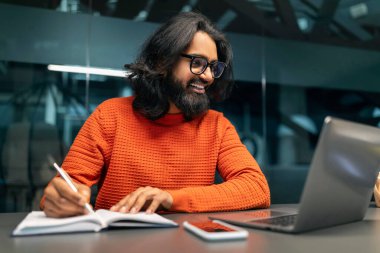 Hintli teknik uzmanlar internetten bilgisayar ekranına bakıyor ve not defterine not alıyor. Sakallı doğulu adamın uluslararası meslektaşlarıyla görüntülü görüşmesi var, modern ofis içi.