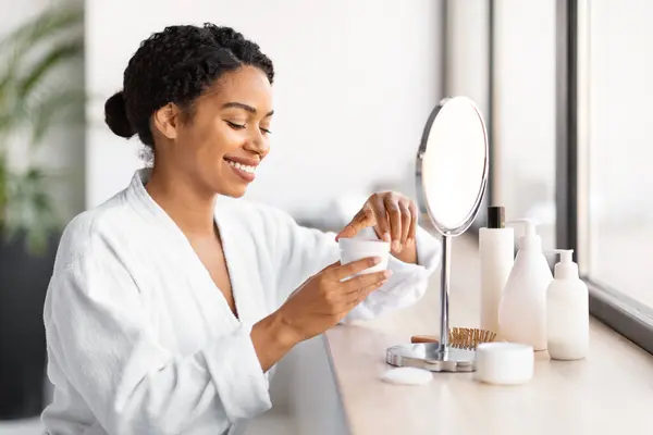 Smiling black woman in white bathrobe opening jar of facial cream, smiling african american female preparing for her skincare routine at home, sitting near mirror in bathroom, closeup shot