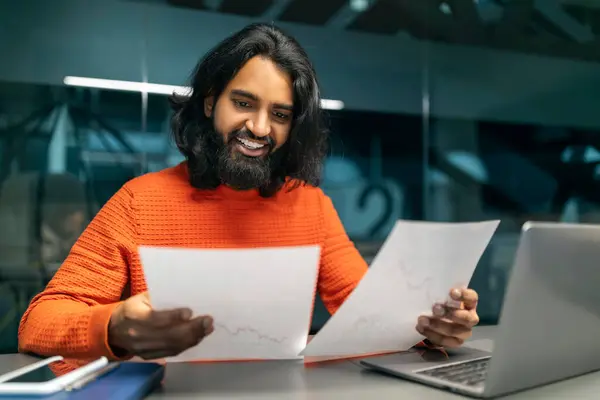 Cheerful millennial bearded long-haired indian businessman doing paperwork at dark office, sitting at desk in front of laptop computer, manager happy with marketing report, copy space