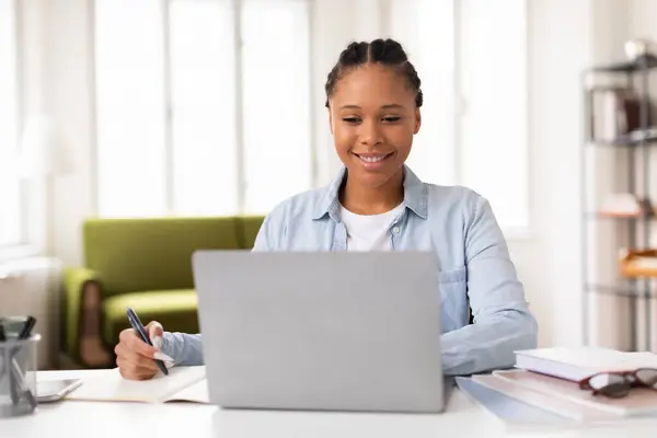 彼女のラップトップを見ながら笑顔でペンを持つ陽気な黒人女性の学生は 自宅で机に座って楽しいオンライン学習経験を示唆しています — ストック写真