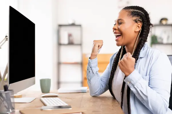 長いブレードを持つ陽気な若い黒人女性の学生は 自宅から勉強して コンピュータとノートブックで彼女の机でエレベーターを感じ 勝利のジェスチャーを作ります — ストック写真