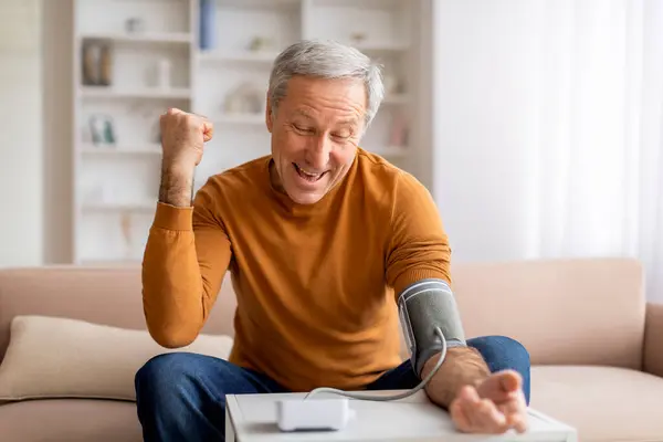 stock image Excited senior man checking blood pressure at home, sitting on couch in living room, looking at electronic tonometer screen and raising hand up, enjoying good checkup results, copy space