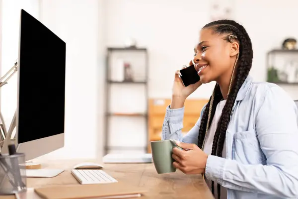 Black lady enjoys warm conversation on her smartphone and holding coffee mug in hand, sitting in front of computer, indicating remote learning from home