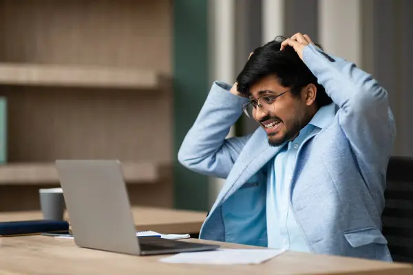 Unhappy Indian professional businessman grasping his head in frustration while staring at his laptop, looking at mirror and mistake in modern office. Workplace stress and panic, business failure
