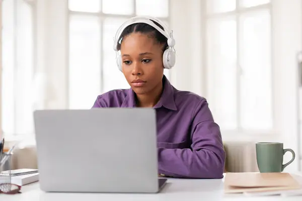 白いヘッドフォンと紫色のシャツを身に着けた黒人女性学生が 明るい部屋に座ってラップトップコンピュータで彼女のオンライン研究に集まった — ストック写真