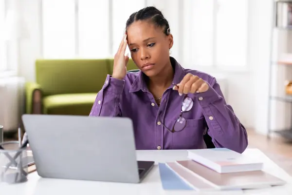 忧心忡忡的黑人女生穿着紫色衬衫 一边在笔记本电脑上学习 一边举着眼镜 一边摸着头 坐在家里的办公桌前 表现出压力的迹象 — 图库照片