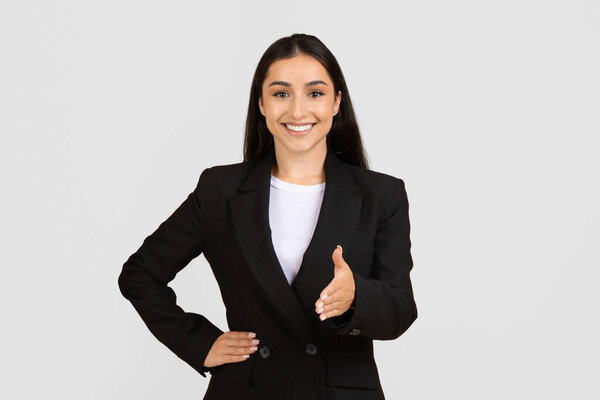Positive young businesswoman in black suit extending her hand for welcoming handshake, posing and smiling at camera on grey background