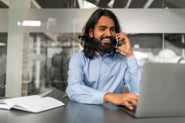Gülümseyen uzun sakallı, uzun saçlı, resmi kıyafet yöneticisi olan, ofisteki masasında oturan, müşterisiyle telefon görüşmesi yapan, dizüstü bilgisayar ekranına bakan, uzayı kopyalayan,