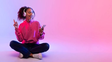 Pembe kapüşonlu ve kotlu neşeli siyahi kadın, beyaz kulaklıkla müzik dinliyor ve akıllı telefon kullanıyor, yerde bağdaş kurarak oturuyor.