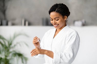 Gülümseyen siyah bir kadın evinde eline nemlendirici serum sürüyor. Beyaz bornozlu çekici bir Afro-Amerikan kadın güzellik rutini yapıyor. Modern banyoda duruyor.