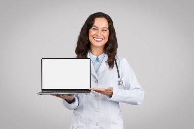 Gülümseyen genç bayan doktor dizüstü bilgisayarı tıbbi içerik için beyaz ekranla sunuyor, tele sağlık reklamları için ideal, gri stüdyo geçmişi, model