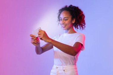 Kıvırcık saçlı neşeli siyah kadın akıllı telefonunu kullanmaya odaklanmış, parlak ışık yüzünü gradyan pembe ve mor arkaplana karşı aydınlatıyor, modern bağlantıyı gösteriyor.