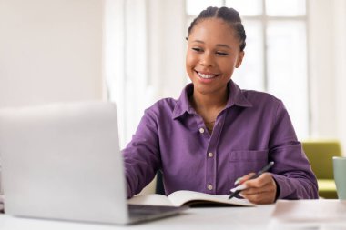 Mor gömlekli neşeli siyahi bayan öğrenci dizüstü bilgisayardaki e-öğrenme seansından zevk alıyor, deftere yazıyor, elinde kalem tutuyor, ışık dolu odada masa başında oturuyor.