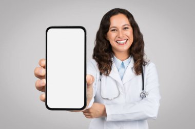 Teletıp teklifi. Genç bayan doktor beyaz ekranlı akıllı telefonu gösteriyor, uygulama veya reklam için sunum yapıyor, gri stüdyo arka planının başında duruyor. Tıp ve mobil teknoloji.
