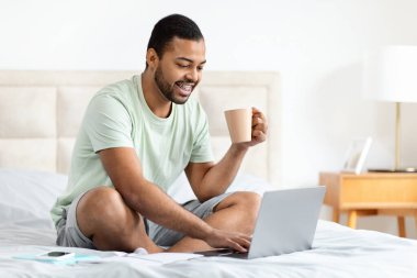 Rahat, genç, siyah bir adam pijama giyiyor, kahve içiyor ve bilgisayar laptopu kullanıyor, evde yatakta oturuyor, web sörfü yapıyor, bilet ayırtıyor, hafta sonu veya tatil planları yapıyor, uzay kopyaları yapıyor.