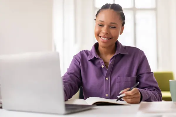 紫色のシャツの陽気な黒人女性学生は ラップトップで彼女のEラーニングセッションを楽しんでいます ノートブックで執筆 ペンを保持 ライト満たされた部屋で机に座って — ストック写真