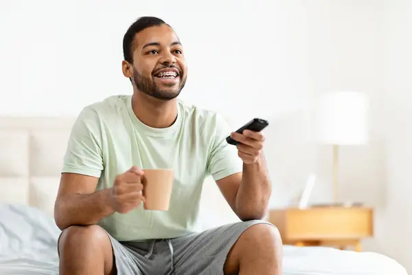 快乐英俊的年轻非洲裔美国男人 穿着睡衣坐在床上 手握遥控器 看电视 看电影或电视剧 喝咖啡 复制空间 — 图库照片