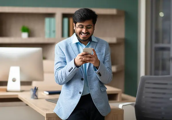 Indian CEO businessman browsing on his smartphone, managing tasks and communication, standing in modern corporate office interior. Success and connectivity in workplace, business applications