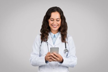 Beyaz üniformalı neşeli Avrupalı bayan doktor, cep telefonu kullanıyor ve gülümsüyor, hastalarla sohbet ediyor, gri arka planda modern sağlık teknolojisini sergiliyor.
