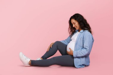 Mutlu genç hamile kadın pembe stüdyo arka planında izole bir şekilde oturuyor, kocaman karnını kucaklıyor ve gülümsüyor, hamileliğin tadını çıkarıyor, bebeğin gelmesini bekliyor, boşluğu kopyalıyor.