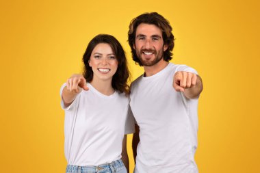Beyaz bluzlu neşeli beyaz bir çift, parlak, çekici bir tebessümle kamerayı işaret ediyor, canlı sarı bir arka plan, stüdyo karşısında birlikte duruyorlar. Motivasyon, reklam ve teklif