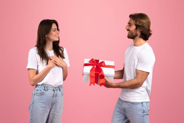 Beyaz bir kutuda kırmızı kurdeleli bir adamla beyaz bir kadını pembe bir geçmişe karşı mutlu bir şekilde sunan mutlu beyaz bir çift, neşe ve kutlama duyguları uyandırıyor.