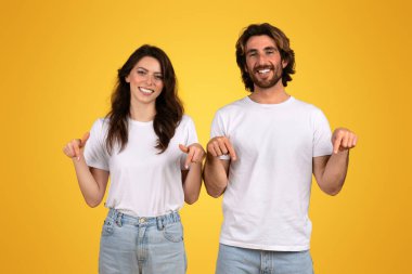 Gülümseyen Avrupalı kadın ve beyaz gömlekli adam, iki elini de aşağı doğru gösteriyor, neşeli ve kendinden emin görünüyor, sarı arka plana karşı duruyor, potansiyel olarak bir ürün veya metin aşağıda