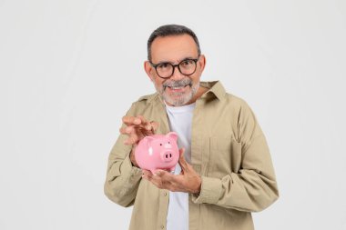 Gülümseyen kıdemli adam pembe kumbaraya bozuk para yerleştiriyor. Zeki tasarruf ve finansal planlamayı temsil eden mutlu yaşlı beyefendi. Stüdyoda beyaz arka planda duruyor.