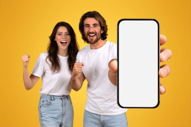 Coşkulu Kafkasyalı genç çift, bir kadının yumruğunu sıkarak başarıyı kutluyor ve bir erkek, sarı arka planda, stüdyoda, boş bir ekranla akıllı telefonu tutuyor.