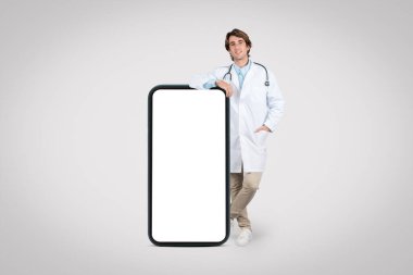 Steteskoplu neşeli genç erkek doktor, laboratuvar önlüğü giyiyor, büyük boy akıllı telefon boş ekranının kenarına yaslanıyor, teletıp