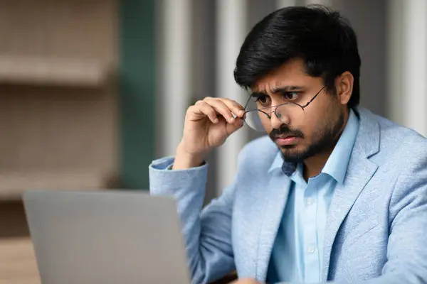 Indian business professional man dealing with eyesight issues, squints at his laptop screen at office. Challenges of managing vision problems in the modern workplace, computer work and eye care