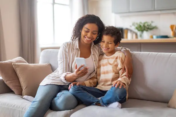 家庭小玩意的概念 快乐的黑人妈妈在智能手机上给她的儿子展示新的应用程序 在网上冲浪或在家里玩网络游戏 同时在舒适的客厅里躺在沙发上休息 — 图库照片