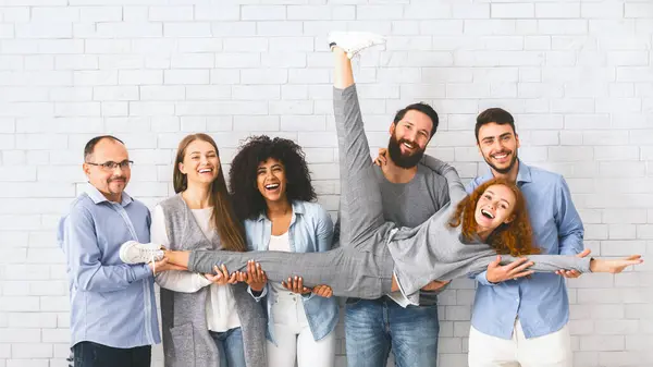 Fun in office. Group of positive people holding their female colleague and laughing while posing over white brick wall background, panorama