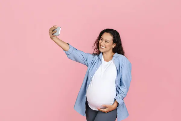 Prenatal and pregnancy blog. Beautiful young pregnant woman wearing casual outfit taking selfie smartphone, expecting influencer streaming, posing isolated on pink studio background