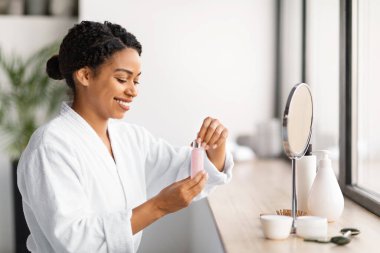 Bornozlu gülümseyen siyah kadın elinde bir şişe yüz serumu, güzel Afrikalı Amerikalı kadın makyaj aynasının yanında oturmuş, kendine bakmanın keyfini çıkarırken cilt bakımı yapmaya hazır.