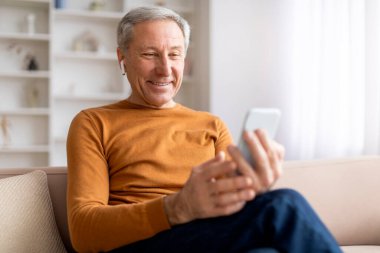 Eğlence, dijital dünya. Evdeki kanepede oturan, cep telefonu ve kablosuz kulaklık kullanan, internetten video yayını izleyen, uzayı kopyalayan mutlu ve yakışıklı bir adam.